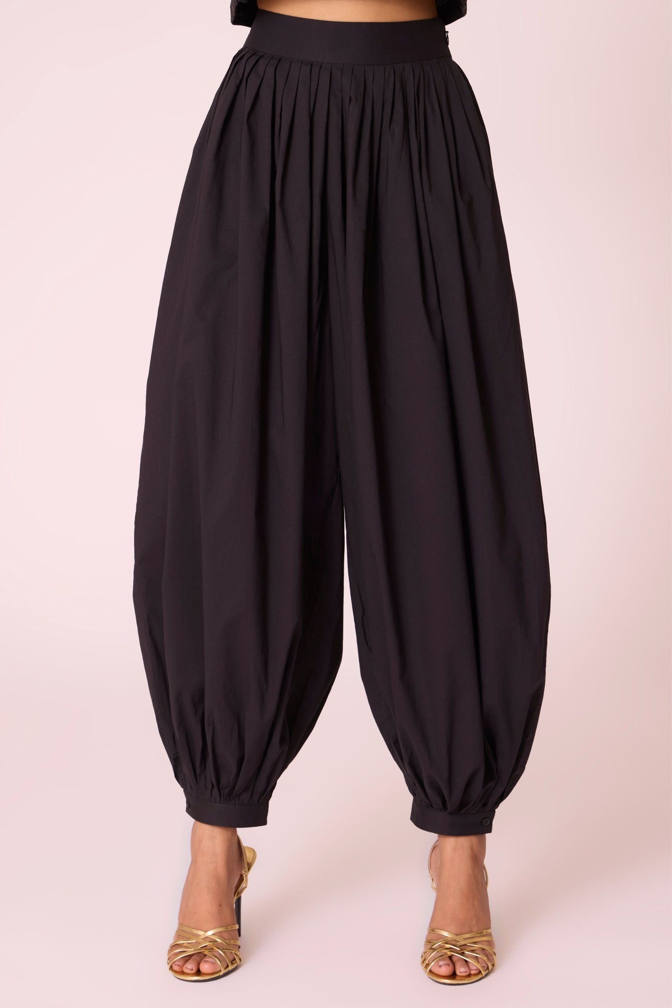 Black organic cotton pleated pants with a relaxed fit, side zip fastening, and adjustable hem cuffs.
